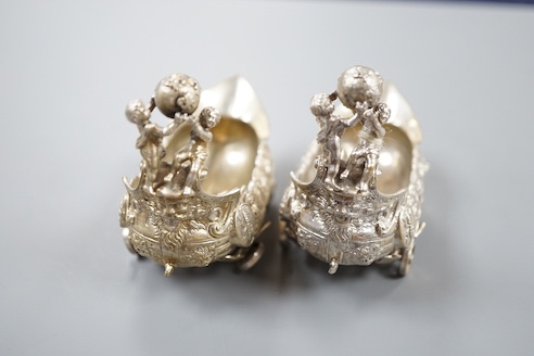 A pair of late 19th/early 20th century Hanau white metal salts, modelled as boat's with wheels and putti surmounts, maker Stork & Sinsheimer, length 81mm, 195 grams.